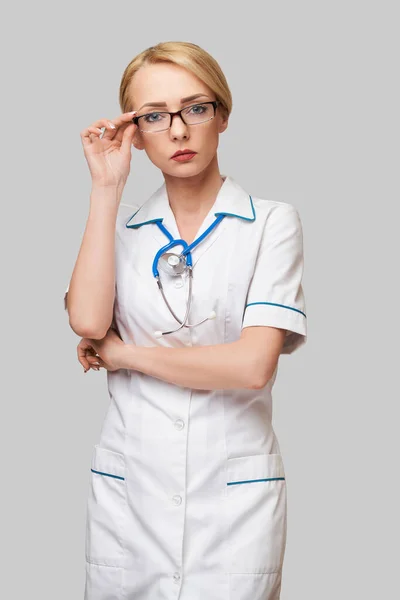 Portrét atraktivní mladé ošetřovatelky v bílém plášti na světle šedém pozadí — Stock fotografie