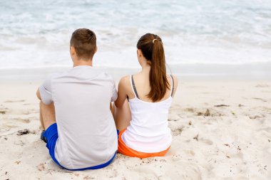 Genç çift sahilde otururken birbirlerine bakıyorlar.