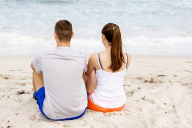 Genç çift sahilde otururken birbirlerine bakıyorlar.