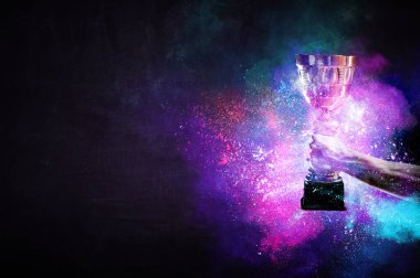 Elinde altın bir kupa var, arka planda.