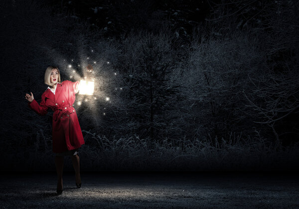 Young blond woman in red cloak with lantern walking in night