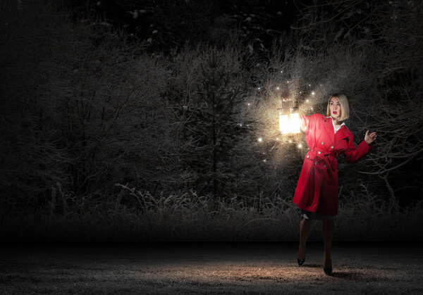 Young blond woman in red cloak with lantern lost in darkness