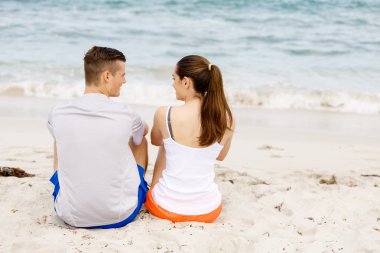 Genç çift sahilde otururken birbirlerine bakıyorlar.