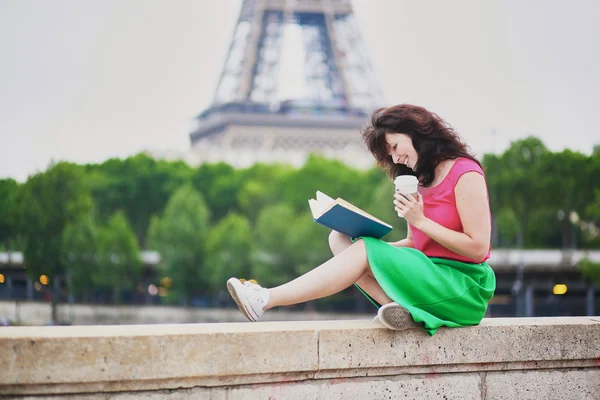 Girl with coffee to go reading a book near the Eiffel tower. — Stock Photo, Image
