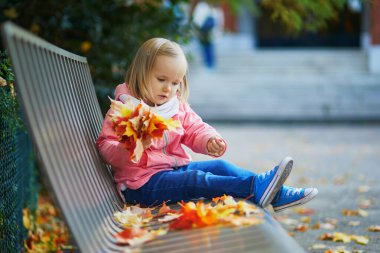 Sevimli küçük kız bankta oturuyor ve sonbahar parkında yaprakları topluyor. Mutlu çocuk Paris, Fransa 'da sonbahar gününün tadını çıkarıyor. Çocuklar için açık hava etkinlikleri