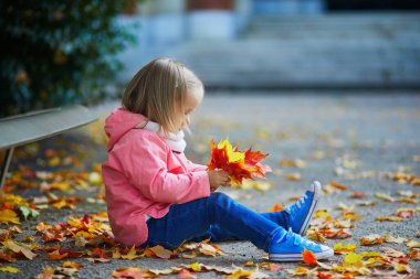Sonbahar parkında yerde oturup düşen yaprakları toplayan sevimli küçük kız. Mutlu çocuk Paris, Fransa 'da sonbahar gününün tadını çıkarıyor. Çocuklar için açık hava etkinlikleri