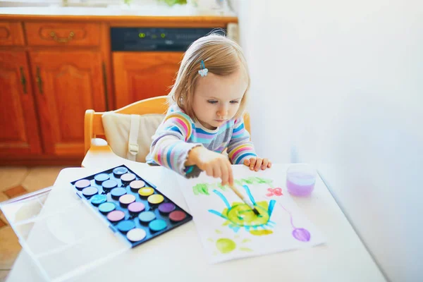 Adorable little girl drawing with colorful aquarelle paints at home, in kindergarten or preschool. Creative games, education and distance learning for kids. Stay at home entertainment