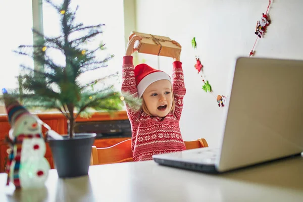 現在とラップトップでサンタの帽子の幸せな幼児の女の子 クリスマスや新年に友人や親戚と通信するためにコンピュータを使用して子供 家にいながらオンラインで季節の休日を祝う — ストック写真