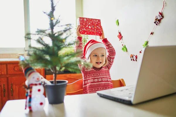現在とラップトップでサンタの帽子の幸せな幼児の女の子 クリスマスや新年に友人や親戚と通信するためにコンピュータを使用して子供 家にいながらオンラインで季節の休日を祝う — ストック写真