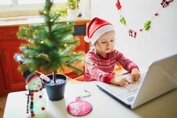 ラップトップとサンタの帽子の幸せな幼児の女の子 クリスマスや新年に友人や高齢者の親戚と通信するためにコンピュータを使用して子供 家にいる間にオンラインで季節の休日を祝う — ストック写真