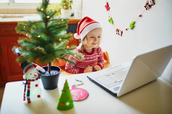 ラップトップとサンタの帽子の幸せな幼児の女の子 クリスマスや新年に友人や高齢者の親戚と通信するためにコンピュータを使用して子供 家にいる間にオンラインで季節の休日を祝う — ストック写真