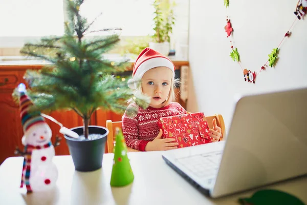 現在とラップトップでサンタの帽子の幸せな幼児の女の子 クリスマスや新年に友人や親戚と通信するためにコンピュータを使用して子供 家にいながらオンラインで季節の休日を祝う — ストック写真