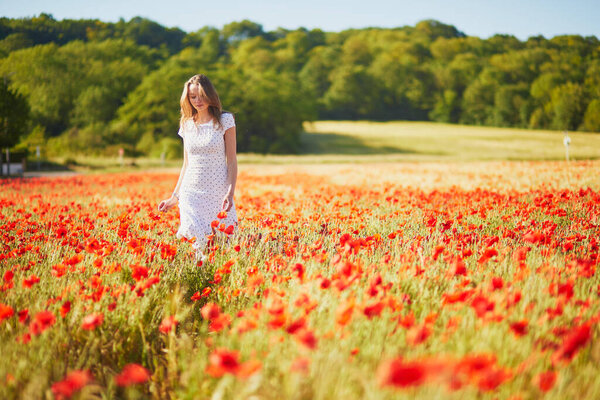 Beautiful young woman in white dress walking in poppy field on a summer day. Girl enjoying flowers in countryside