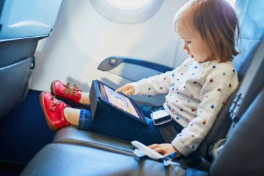Küçük sevimli kız uçakla seyahat ediyor. Küçük bir çocuk uçak penceresinin yanında oturuyor ve uçuş sırasında dijital tablet kullanıyor. Çocuklarla yurtdışına seyahat etmek. Refakatsiz küçük konsept
