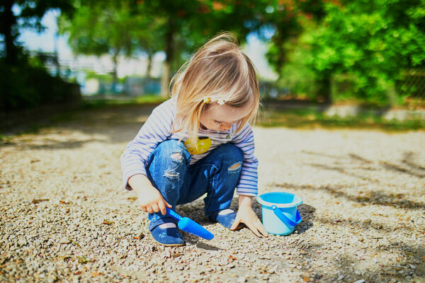 Очаровательная девочка играет с ведром и лопатой, делает грязь и собирает маленькие камни. Активный отдых на свежем воздухе для детей