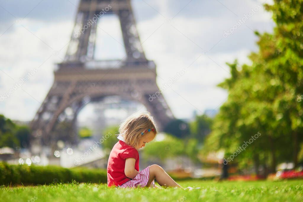 Unhappy and gloomy toddler girl sitting on the grass near the Eiffel tower in Paris, France