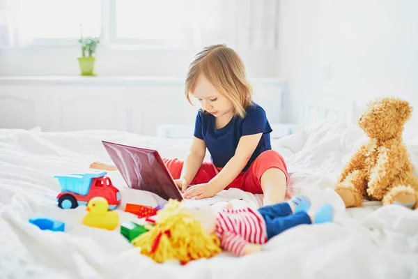 ラップトップ ノートブック きれいな白いリネンのベッドの中で異なるおもちゃを持つ幼児の女の子 フリーランス 遠隔教育 または子供の概念を持つ家庭からの仕事 — ストック写真