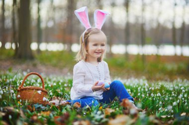 Tavşan kulaklı bir kız Paskalya 'da yumurta avı oynuyor. Bir sürü kartopu çiçeğiyle çimenlerde oturan ve renkli yumurtaları sepette toplayan bir çocuk. Paskalyayı dışarıda kutlayan küçük çocuk.