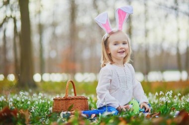 Tavşan kulaklı bir kız Paskalya 'da yumurta avı oynuyor. Bir sürü kartopu çiçeğiyle çimenlerde oturan ve renkli yumurtaları sepette toplayan bir çocuk. Paskalyayı dışarıda kutlayan küçük çocuk.