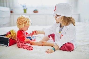 Beyaz önlüklü, şırıngayla doktorculuk oynayan ve bebeğine tıbbi bakım yapan sevimli bir kız. Çocuklar ve rol oyunları. Covid-19 aşı kavramı