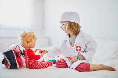 Beyaz önlüklü, şırıngayla doktorculuk oynayan ve bebeğine tıbbi bakım yapan sevimli bir kız. Çocuklar ve rol oyunları. Covid-19 aşı kavramı