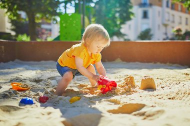 Sevimli küçük kız kum havuzunda oyun parkında eğleniyor. Bebek kum kalıplarıyla oynuyor ve çamur yapıyor. Çocuklar için açık hava yaratıcı aktiviteler