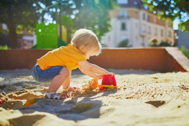 Sevimli küçük kız kum havuzunda oyun parkında eğleniyor. Bebek kum kalıplarıyla oynuyor ve çamur yapıyor. Çocuklar için açık hava yaratıcı aktiviteler
