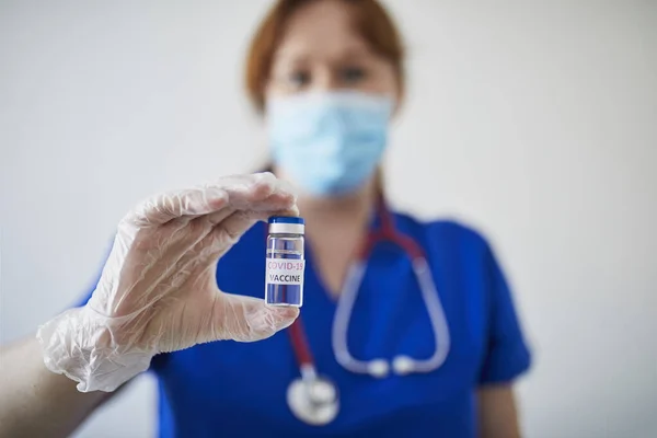 Female doctor or nurse with stethoscope, wearing face mask and sterile gloves, holding vial with COVID-19 vaccine. Research, clinical trial, vaccination or herd immunity concept