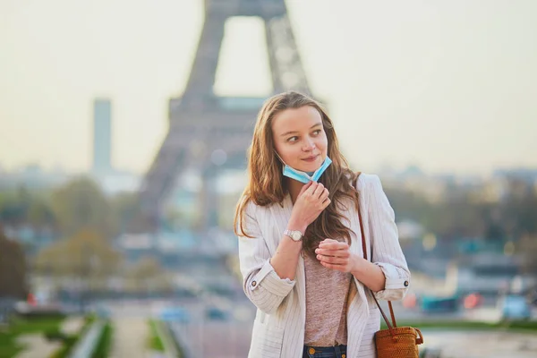 Young girl standing near the Eiffel tower in Paris and removing protective face mask during coronavirus outbreak. Pandemic and lockdown in France. Tourist spending vacation in France during pandemic