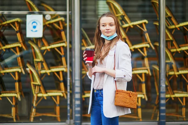 Young girl standing near closed restaurant in Paris and wearing protective face mask during coronavirus outbreak. Pandemic and lockdown in France. Tourist spending vacation in France during pandemic