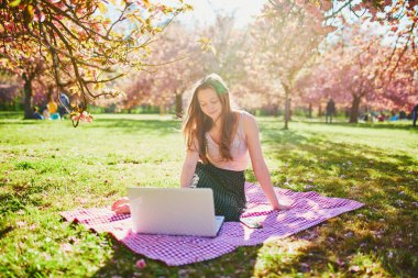 Güzel genç kız kiraz çiçeği mevsiminde parkta dizüstü bilgisayarıyla çalışıyor. Paris, Fransa yakınlarındaki ünlü Sceaux Parkı 'nda genç bir kadın. Serbest çalışma, uzaklık öğrenme veya uzaktan çalışma kavramı