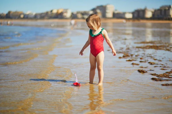 在法国布列塔尼的圣马洛海滩上 穿着红色游泳衣的可爱的幼儿小女孩正在玩水中的木制玩具船 孩子们的户外暑期活动 在海上度假的孩子 — 图库照片