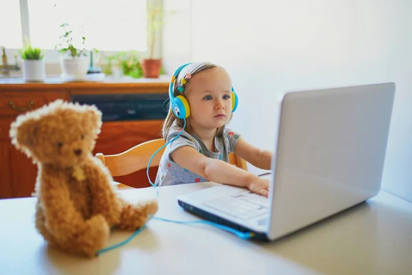 Toddler girl wearing eraphones with laptop. Kid using computer to communicate with friends, elderly relatives or kindergartners. Education or online communication for kids. Stay at home entertainment