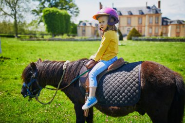 Üç yaşındaki sevimli bir kız midilliye biniyor. Küçük çocuk ata binme alıştırması yapıyor. Küçük çocuklar için binicilik. Çocuklar için açık hava yaz etkinlikleri