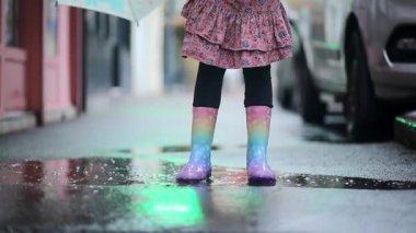 Yağmurlu bir günde su birikintisinde atlarken gökkuşağı lastik çizmeleri ve pembe bir elbise giyen genç bir kızın yakın çekimi. Eğlenceli çocukluk aktiviteleri, yağmurlu hava ve sokak yansımaları..