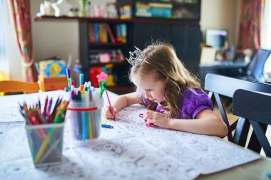 Evde kalemler ve kalemlerle bir masada prenses elbisesi boyayan küçük bir kız. Sanat malzemeleri ve boyama kağıtlarıyla yaratıcı iç etkinlik