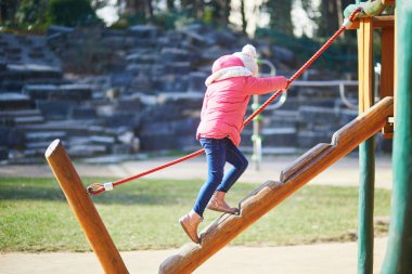 Sıcak kış kıyafetleri içindeki genç bir kız dışarıdaki tahta oyun parkına tırmanıyor ve dengede duruyor. Aktif çocukluk anı hareket, koordinasyon ve açık hava oyunlarına odaklandı