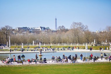 Paris, Fransa - 17 Mart 2025: Güneşli bir bahar gününde Paris 'teki Jardin du Lüksemburg' daki merkez çeşmenin etrafında dinlenen kalabalık, arka planda Eyfel Kulesi ile birlikte.