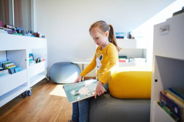 Bir halk kütüphanesinin parlak çocuk bölümünde otururken renkli bir resim kitabı okuyan genç bir kız. Eğitici ve huzurlu çocukluk dönemi öğrenme ve merak.