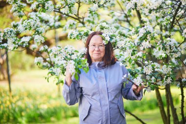 Gözlüklü ve açık mavi ceketli yaşlı kadın bir bahar bahçesinde çiçek açan bir ağacın dalları arasında duruyor..