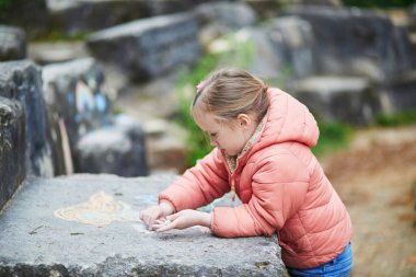 Pembe ceketli küçük kız dışarıdaki büyük bir taş levhaya tebeşirle renkli şekiller çiziyor..