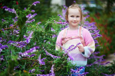 Pastel bir süveter ve sırt çantası giyen neşeli genç kız çiçek açan bahçedeki yemyeşil mor çiçeklerin arasında duruyor..