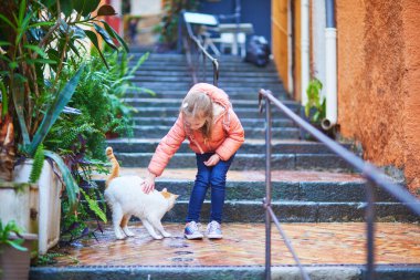 Turuncu ceketli genç kız dışarıda taş merdivenlerde çömelmiş beyaz ve turuncu bir kediyi okşuyor..