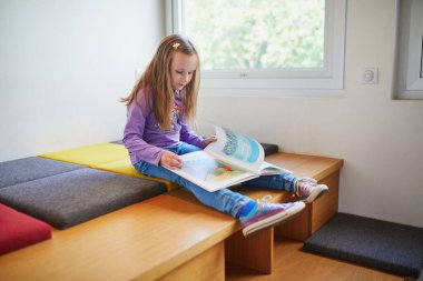 Bir halk kütüphanesinin parlak çocuk bölümünde otururken renkli bir resim kitabı okuyan genç bir kız. Eğitici ve huzurlu çocukluk dönemi öğrenme ve merak.