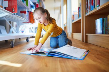 Meraklı genç kız parlak bir kütüphanenin ahşap zemininde diz çökerken renkli bir resim kitabı okuyor. Eğitici ve açık sözlü çocukluk öğrenme ve keşif anı.