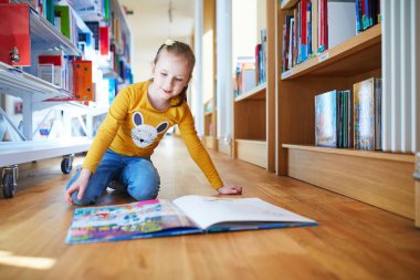 Meraklı genç kız parlak bir kütüphanenin ahşap zemininde diz çökerken renkli bir resim kitabı okuyor. Eğitici ve açık sözlü çocukluk öğrenme ve keşif anı.