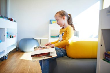 Bir halk kütüphanesinin parlak çocuk bölümünde otururken renkli bir resim kitabı okuyan genç bir kız. Eğitici ve huzurlu çocukluk dönemi öğrenme ve merak.