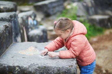 Pembe ceketli küçük kız dışarıdaki büyük bir taş levhaya tebeşirle renkli şekiller çiziyor..