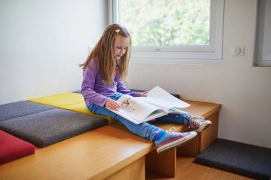 Bir halk kütüphanesinin parlak çocuk bölümünde otururken renkli bir resim kitabı okuyan genç bir kız. Eğitici ve huzurlu çocukluk dönemi öğrenme ve merak.