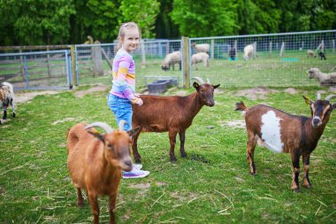 Evcil hayvan ya da çiftlikte keçiye bakarken çömelmiş gülümseyen küçük kız..